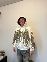 Hoodie oversize de color blanco con estampado artístico en tonos marrones y dorados en el pecho y las mangas. Presenta un forro interior de capucha en tejido marrón texturizado y costuras reforzadas.