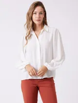 Camisa de gasa blanca con cuello solapa, cierre frontal con botones y detalle de puntilla a lo largo de la abertura. Presenta mangas largas con puños elásticos y costuras decorativas en el antebrazo.