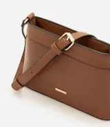 Cartera bandolera pequeña color beige con ribetes marrones, de la marca Satinato. Presenta cierre de cremallera, asa larga ajustable y detalle de placa metálica dorada con el logo en el frente.