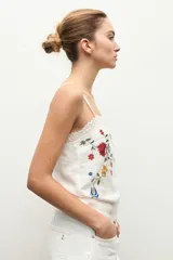 Top blanco de tirantes finos con detalle de encaje en escote y dobladillo, con bordado floral multicolor en el frente.