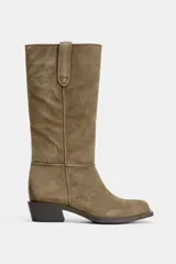 Bota de caña alta de serraje color verde oliva, estilo cowboy, con tiradores en la caña y pespuntes visibles. Presenta un tacón bajo y ancho de bloque y punta cuadrada.