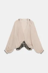Blusa color beige confeccionada en viscosa, con cuello redondo y escote pico. Tiene mangas largas con puños y detalle de bordados con aplicación de abalorios en cuello, puños y ruedo. Diseño cruzado con fajín.