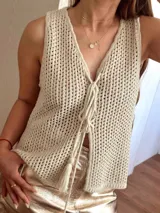 Chaleco tejido en punto calado con hilo de algodón y lurex, de color negro con destellos plateados. Presenta escote profundo en V, cierre frontal con cordones entrelazados terminados en borlas y un corte recto y holgado.