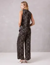 Conjunto de dos piezas compuesto por una blusa sin mangas con cuello solapa y escote en V con botones frontales, y un pantalón de tiro alto con pierna ancha y bolsillos laterales. Ambas prendas están confeccionadas en satén con un estampado floral sobre fondo oscuro.