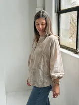 Camisa de lino color beige con diseño oversize, cuello camisero mao, cierre frontal con botones ocultos, mangas largas abullonadas con puños elásticos y delicados bordados florales tono sobre tono en el frente y las mangas.