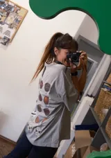 Remera oversize de algodón en color gris, con escote bote y costuras a la vista. Presenta un diseño estampado en la espalda con múltiples círculos que ilustran logros de la selección uruguaya y un parche decorativo.