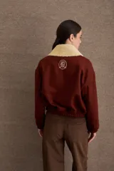 Campera bomber marrón de paño de lana, con cierre frontal, cuello de corderito sintético blanco desmontable, bolsillos laterales, puños y dobladillo elásticos, y logo bordado en el pecho.