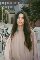 Vestido corto de tejido fluido en color beige rosado, con cuello subido ajustable mediante lazada en la espalda. Presenta mangas largas abullonadas con puños abotonados, cintura entallada y falda con bajo acabado en volantes. Incluye forro interior y cierre lateral con cremallera oculta.