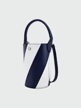 Cartera tipo bucket mini de cuero, con diseño bicolor en franjas diagonales azul marino y blanco. Cuenta con un asa superior rígida, correa ajustable para el hombro, cierre superior y detalle de botón metálico con logo grabado.