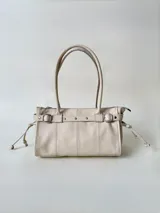 Cartera de cuero de tamaño medio con estructura rígida. Presenta una tira decorativa con tachas y hebillas metálicas en los extremos, además de lazos laterales. Cuenta con doble asa para llevar al hombro y cierre superior.