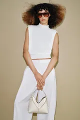 Bolso tipo mini shopper de diseño estructurado en color beige, con forma de V en la parte superior y asa de mano fija. Presenta costuras visibles y detalles metálicos en los laterales.
