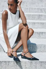 Sandalia negra de charol con punta fina, taco kitten y pulsera ajustable al tobillo. Presenta una tira delantera con hebilla dorada.