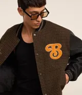 Campera bomber de estilo college con cuerpo a cuadros en tonos marrones y mangas largas de cuero sintético negro. Presenta cuello, puños y cintura acanalados a rayas, cierre abotonado, bolsillos laterales y aplique bordado de letra "B" en el pecho.