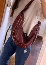 Cartera estilo hobo de cuero sintético color bordó, con diseño de media luna y detalles de ojales metálicos plateados a lo largo de los bordes. Cuenta con correa ajustable y cierre superior.