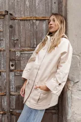 Parka liviana color beige con capucha, cierre frontal con botones, bolsillos delanteros con tapa y ajuste en la cintura. Presenta mangas con puños vueltos y detalle de botones.