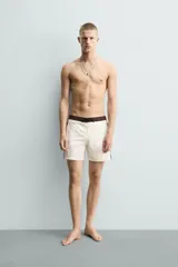 Short de baño blanco marfil con cintura marrón y cordón ajustable del mismo color. Presenta franjas laterales en tono azul marino con finos bordes rojizos y bolsillos laterales.