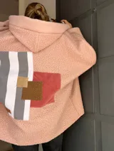 Campera de corderito sintético color rosa, con capucha y diseño de collage en la espalda que combina franjas verticales en tonos grises, blanco y parches en texturas de marrón y terracota. Presenta un corte holgado y mangas anchas.