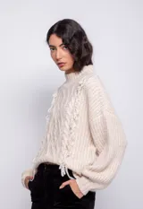 Sweater color crema de tejido trenzado con cuello alto, mangas largas abullonadas y detalle de flecos en el frente.