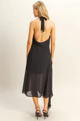 Vestido largo negro con escote halter y espalda descubierta. Presenta un diseño asimétrico con una capa superpuesta de tela semitransparente que cae lateralmente, creando un efecto de movimiento y elegancia.