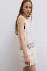 Vestido corto de gasa color blanco, con escote en V cruzado, sin mangas y diseño con volantes en el bajo. Presenta un detalle de tachas plateadas en la cintura.