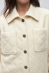 Camisa de lana fina sin teñir, color marrón, con diseño capitoneado y pespuntes decorativos. Presenta cuello clásico, cierre frontal con botones metálicos y dos bolsillos frontales tipo parche.