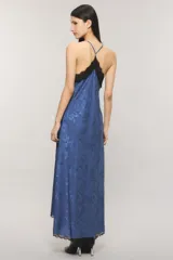 Vestido maxi azul de satén con diseño levemente asimétrico, corte al bies y escote en V con detalle de puntilla negra. Presenta espalda deportiva con tirantes finos regulables y ribete de encaje en el ruedo.