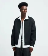 Campera masculina de corte boxy, color negro, con cuello inglés, cierre frontal con cremallera, mangas largas con puños elásticos y ruedo con elástico. Presenta detalles de recortes en el diseño.