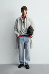 Trench coat de corte relajado, color gris claro, confeccionado en tejido técnico con mezcla de algodón. Presenta cuello solapa, manga larga con puño ajustable, bolsillos de doble vivo en cadera, bolsillo interior y abertura en el centro de la espalda. Cierre frontal con botonadura oculta.