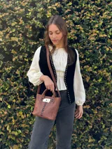 Cartera de cuero marrón de formato estructurado, con doble asa de mano y correa larga ajustable para llevar al hombro o cruzada. Cuenta con detalles de costuras negras a la vista y una pieza decorativa central de resina con efecto carey y aplique metálico.