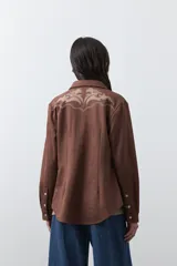 Sobrecamisa de gamuza color marrón con bordados decorativos estilo western en la zona de los hombros y cierre frontal con botones metálicos.