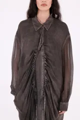 Vestido camisero largo midi de silueta envolvente, confeccionado en tejido liviano y fluido, color gris oscuro con efecto desgastado. Presenta cierre frontal con botones ocultos, cuello clásico, mangas largas con puño y frunces centrales que aportan volumen y movimiento.