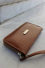 Billetera tipo sobre con correa de muñeca, confeccionada en cuero con textura de reptil en color marrón. Cuenta con cierre perimetral y placa metálica con el logo de la marca en el frente.