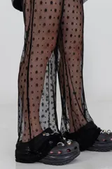 Pantalón acampanado de encaje negro, con estampado de pequeñas flores en relieve. El tejido es semitransparente y cae sobre un calzado tipo zueco negro.