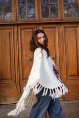 Poncho tejido de color blanco, con flecos en los bordes y detalles de bordado en crochet a lo largo de la abertura frontal. Cuenta con un cierre de cordones con borlas en el cuello.
