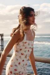 Vestido mini de color beige con un estampado de tomates, aceitunas y botellas de aceite, y un borde inferior rojo con ilustraciones de una sombrilla de playa y vegetales. Cuenta con escote cuadrado y tirantes anchos cruzados en la espalda.