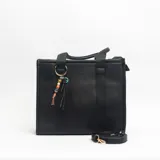Cartera tipo tote de estructura rígida y diseño minimalista en color negro. Presenta doble asa de mano y una correa larga ajustable y extraíble. Incluye un detalle decorativo de argolla metálica con cuentas de colores y tiras de cuero sintético colgantes.