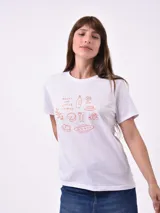 Remera blanca de manga corta con cuello redondo y un estampado frontal en color terracota que incluye ilustraciones de alimentos (botella, copa, pan, queso, etc.) y texto que dice "Enjoy the little things" y "Things between".