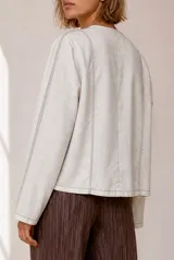 Chaqueta de lino color beige, de corte recto, escote redondo y cierre frontal con broches a presión. Cuenta con mangas largas y costuras decorativas pespunteadas en el frente.