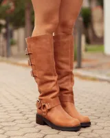 Bota de caña alta confeccionada en cuero color marrón tostado. Presenta un diseño con tres hebillas decorativas en la parte superior de la caña y una correa con hebilla y argolla metálica en el tobillo. Cuenta con suela plana y taco bajo de madera oscura.