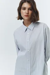 Camisa de algodón con patrón de rayas verticales finas en tonos celeste pálido y blanco. Presenta un corte oversized, cuello clásico y puños abotonados.