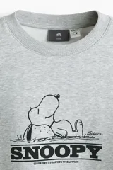 Buzo gris melange de corte holgado, con hombros caídos, cuello redondo, puños y dobladillo acanalados. Cuenta con un estampado frontal de Snoopy en el centro del pecho.