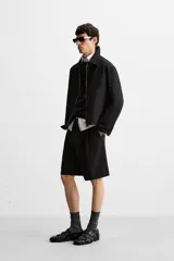 Bermuda de corte relajado color negro, con pliegues frontales, cierre de cremallera y botón, bolsillos laterales y bolsillos traseros.