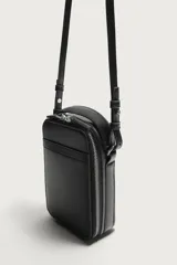 Bandolera vertical lisa de color negro, con compartimento principal con cierre de cremallera, bolsillo interior plano y asa de hombro ajustable.