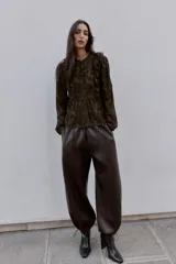 Blusa de manga larga con cuello redondo, confeccionada en tejido semitransparente con estampado paisley en tonos marrones oscuros. Presenta detalles de volantes y apliques de terciopelo en el frente y hombros, con cierre frontal de botones forrados.