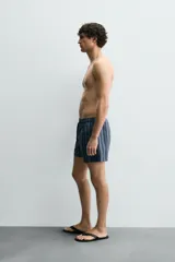 Bañador de hombre de largo medio, color azul claro con rayas verticales en azul oscuro. Posee cintura elástica ajustable con cordón, bolsillos laterales y un bolsillo trasero de plastrón.