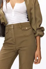 Pantalón de sarga color tostado, de tiro alto y corte acampanado con diseño tobillero. Presenta bolsillos frontales tipo parche con solapa abotonada y pliegues marcados al frente y en la espalda.