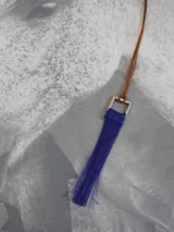 Collar con cordón de cuero marrón y dije cuadrado metálico plateado del cual cuelga una borla de hilos de seda en color azul.