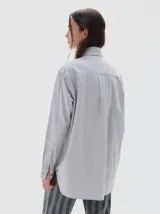 Camisa oversized de algodón color gris claro, con cuello clásico, botones negros y bolsillo frontal con etiqueta de marca.