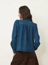 Camisa de jean de corte recto con cuello alto tipo mao con volados, cierre frontal con botones a presión y mangas largas con hombros ligeramente abullonados.