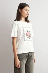 Remera color crudo de manga corta, con cuello redondo y un aplique bordado frontal con diseño floral enmarcado por pespuntes.